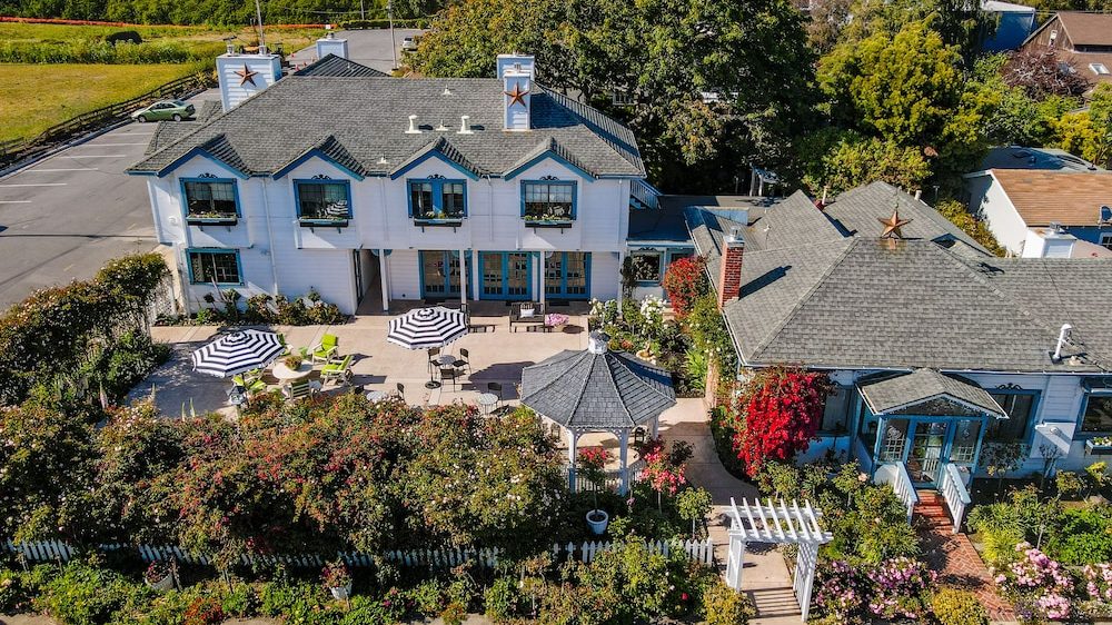 Two connected white houses with gray shingled roofs, decorated with large star ornaments on the chimneys. The front yard features a patio with striped umbrellas, green and beige chairs, and a white gazebo surrounded by lush greenery and colorful flowering plants. A white picket fence and a white arbor gate line the sidewalk in front. Trees and a parking lot are visible in the background.