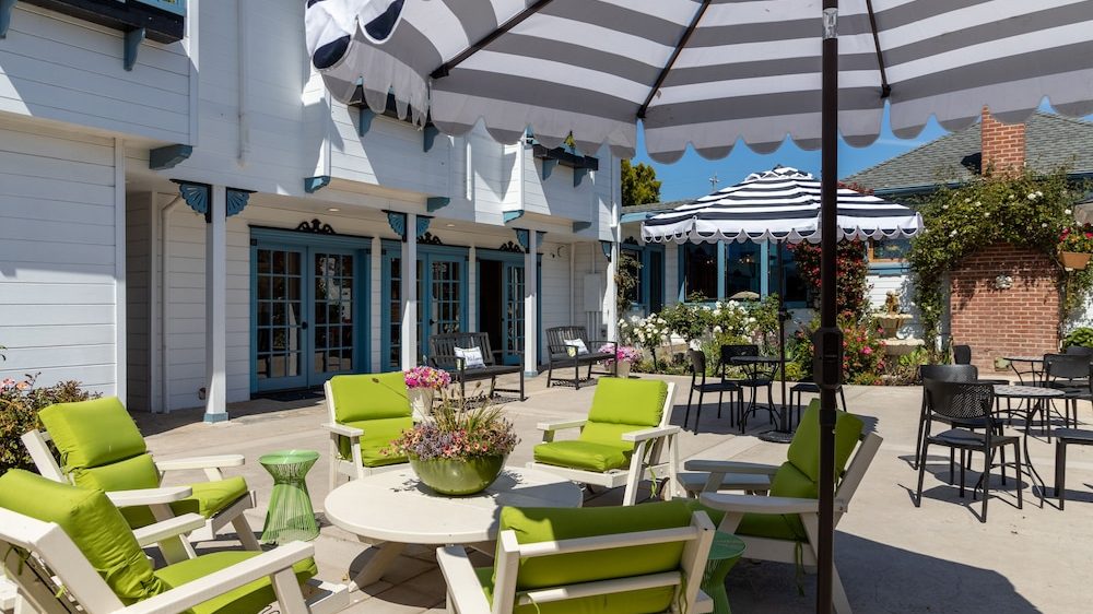 Outdoor patio area with white wooden chairs featuring bright green cushions arranged around a round white table with a flower centerpiece. Two large black and white striped umbrellas provide shade. The background includes a white building with blue-trimmed windows and doors, and a garden with blooming flowers and greenery.
