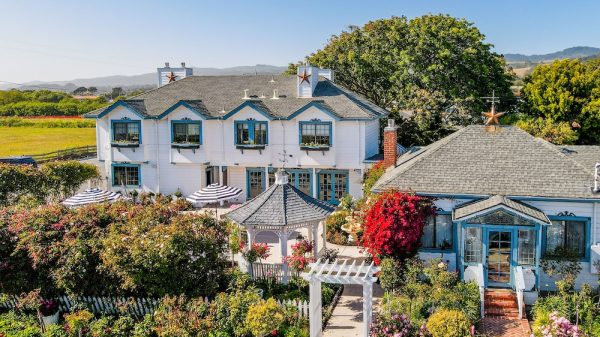 Two white houses with blue trim and gray shingled roofs are surrounded by lush gardens filled with colorful flowers and greenery. A white gazebo with a gray roof sits between the houses, and a white pergola marks the entrance to the garden path. The scene is set under a clear blue sky with trees and hills in the background.
