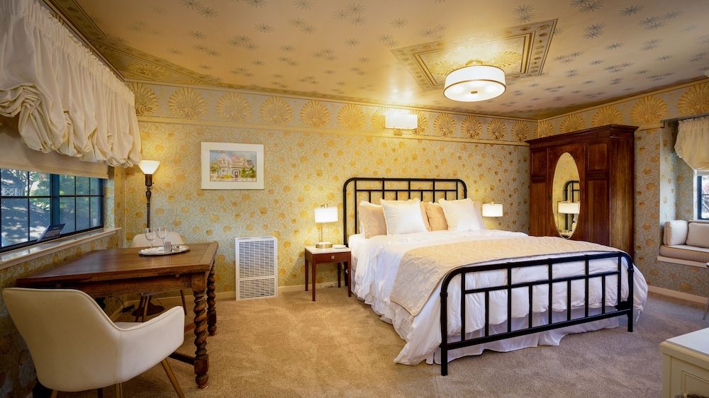Bedroom with a black metal bed frame, white bedding, and beige pillows. The room features patterned wallpaper with gold accents, a wooden wardrobe with an oval mirror, a wooden desk with a white chair, and a window with a cushioned bench. The ceiling has a decorative light fixture and matching wallpaper pattern. The floor is carpeted in beige.