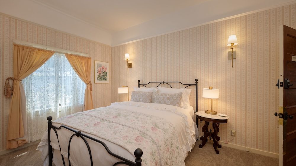 A cozy bedroom with a black metal bed frame, white bedding, and a floral quilt. The walls are covered in light pink patterned wallpaper. There are two wall-mounted lamps above the bed and a small round wooden side table with a lamp on the right side. A window with sheer lace curtains and peach-colored drapes is on the left side of the room. The floor is carpeted in a neutral tone.