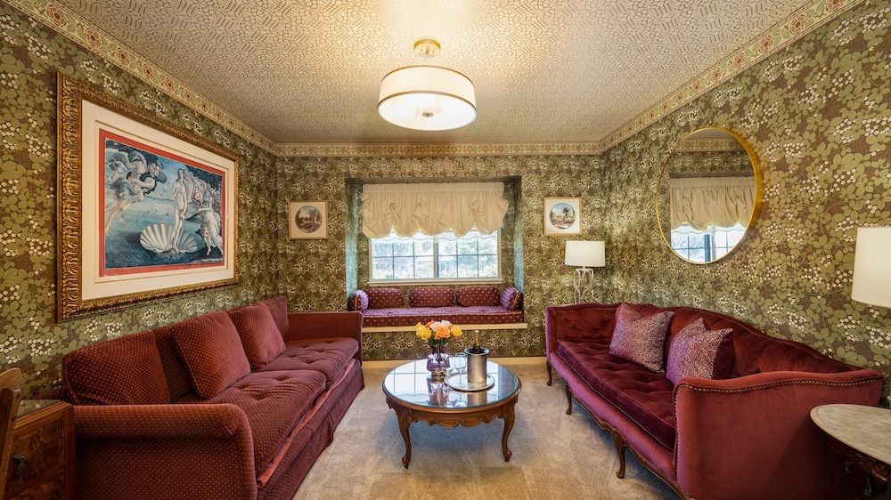 A vintage-style living room with two burgundy velvet tufted sofas facing each other, a round wooden coffee table with a glass top in the center, and a window seat with matching burgundy cushions beneath a window with cream-colored valances. The walls are covered in green floral wallpaper with a decorative border near the ceiling, which has an intricate patterned design. A large framed painting hangs on the left wall, and a round gold-framed mirror is on the right wall. Two small framed pictures flank the window, and there are two table lamps with white shades on either side of the room. The floor is carpeted in a light beige color.