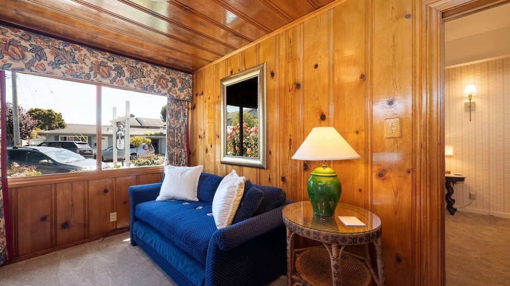 Small cozy room with wood-paneled walls and ceiling, featuring a blue upholstered loveseat with two white pillows, a wicker side table with a green ceramic lamp, a large window with floral valance and curtains, and a framed mirror reflecting flowers outside.