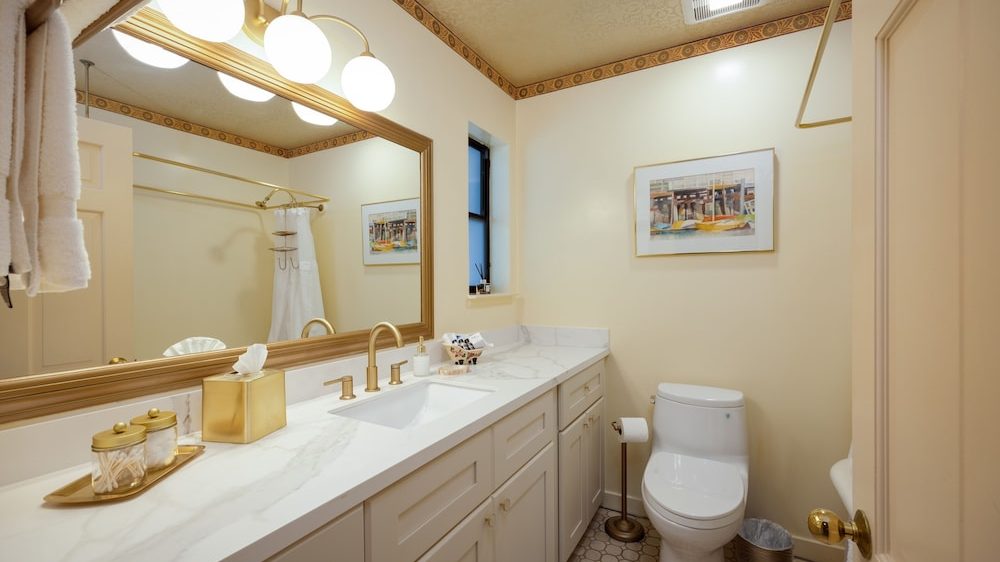 Bathroom with a long white marble countertop featuring a built-in sink and gold faucet fixtures. Above the countertop is a large rectangular mirror with a gold frame, illuminated by three round globe lights. On the countertop are gold-accented containers, a tissue box, and a small basket with toiletries. The walls are painted a light cream color with a decorative border near the ceiling. A white toilet is positioned next to the countertop, with a gold toilet paper holder and a small trash can nearby. A framed picture hangs on the wall above the toilet, and a small window is set into the wall above the countertop. The floor has a hexagonal tile pattern in neutral tones.