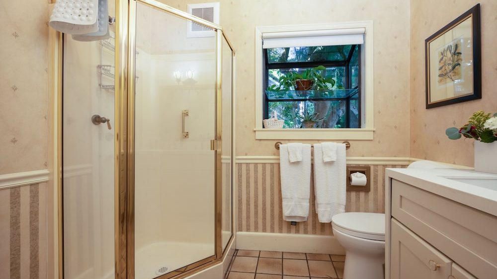 Bathroom with a corner glass shower enclosure featuring gold trim, a white toilet, a vanity with a white countertop, and a window with plants on the sill. Two white towels hang on a towel rack beneath the window, and a framed botanical print is on the wall above the toilet. The walls have a beige wallpaper with a striped lower half, and the floor is tiled in a light brown color.