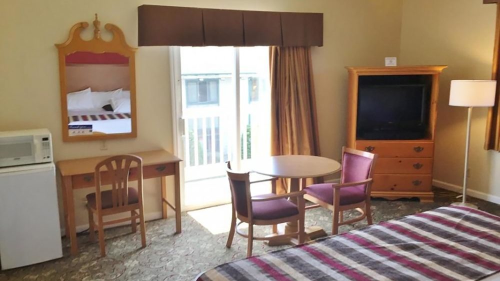 Hotel room with a bed covered by a striped blanket in shades of red, gray, and beige. There is a small wooden table with two purple cushioned chairs near a sliding glass door with brown curtains. A wooden dresser with a TV on top and a floor lamp beside it are visible. On the left side, there is a wooden desk with a chair, a microwave on top of a mini fridge, and a decorative mirror above the desk. The room has beige walls and patterned carpet flooring.