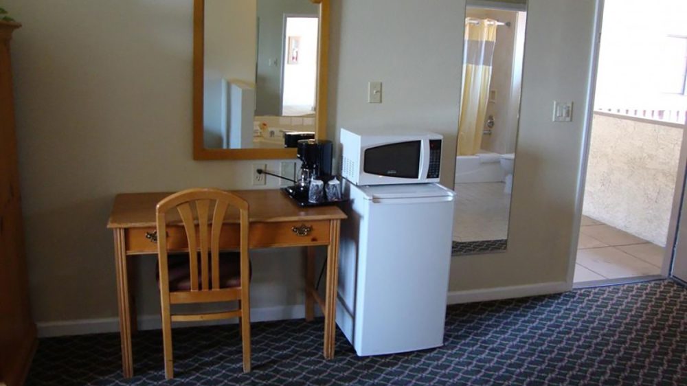 Wooden desk with a matching chair, a coffee maker, and two coffee cups on top. Above the desk is a decorative wooden-framed mirror. Next to the desk is a white mini refrigerator with a microwave on top. A full-length mirror is mounted on the wall beside the refrigerator, reflecting a bathroom with a bathtub and shower curtain. The floor is covered with a dark patterned carpet, and there is an open door leading to a tiled outdoor area.