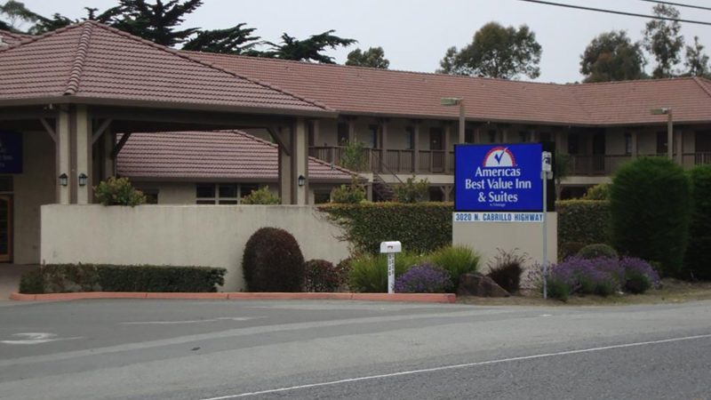 Americas Best Value Inn & Suites building with a brown tiled roof, beige walls, and a blue sign displaying the hotel name and address "3020 N. Cabrillo Highway." The area around the sign has green bushes and purple flowers, and there is a paved road in the foreground.