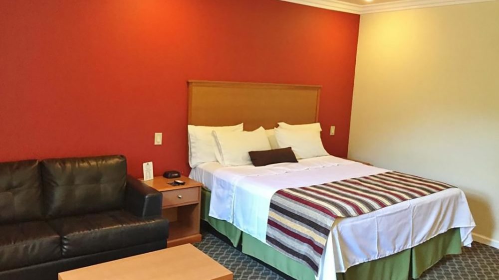 Hotel room with a large bed featuring white sheets, multiple white pillows, and a striped blanket. The bed has a wooden headboard and a green bed skirt. To the left of the bed is a wooden nightstand with a phone and a small card. Next to the nightstand is a black leather couch. In front of the couch is a simple wooden coffee table. The walls are painted red behind the bed and beige on the adjacent wall, with white crown molding along the ceiling. The floor is carpeted with a patterned design.