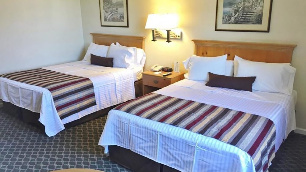 Two double beds with wooden headboards, white linens, striped bed runners, and dark brown accent pillows in a hotel room. A wooden nightstand with a telephone and alarm clock sits between the beds, beneath a wall-mounted light fixture. Two framed landscape paintings hang above the beds, and the floor is carpeted with a patterned design. A round wooden table is partially visible in the foreground.