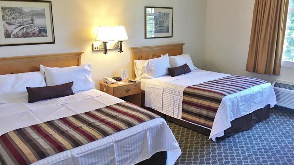 Two double beds with wooden headboards, white linens, striped bed runners in shades of brown, beige, and burgundy, and dark brown accent pillows. A wooden nightstand with a telephone, alarm clock, and remote controls sits between the beds. A wall-mounted light fixture is above the nightstand, and two framed pictures hang on the wall. Beige walls, patterned carpet, and a window with brown curtains are visible.