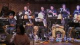 A jazz band is performing on stage with a brick wall background. The band members, mostly older men, are seated and playing various instruments including saxophones, clarinets, and a double bass. A grand piano is visible on the left side. In the foreground, a woman in a striped shirt is smiling and reaching out, partially obscuring the view of the band. The stage is lit with warm lighting.