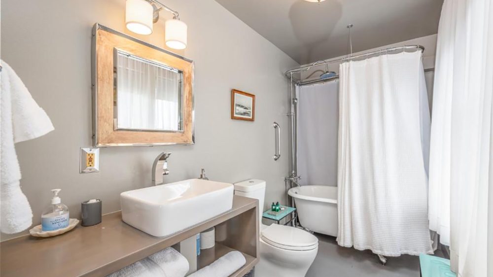 Modern bathroom with a rectangular white vessel sink on a taupe countertop, a large square mirror with a metallic frame above it, and two wall-mounted lights. The room features a white toilet, a clawfoot bathtub with a white shower curtain, and a small glass side table holding toiletries. White towels are neatly stacked on an open shelf under the countertop. The walls and floor are light gray, and sheer white curtains cover the window.