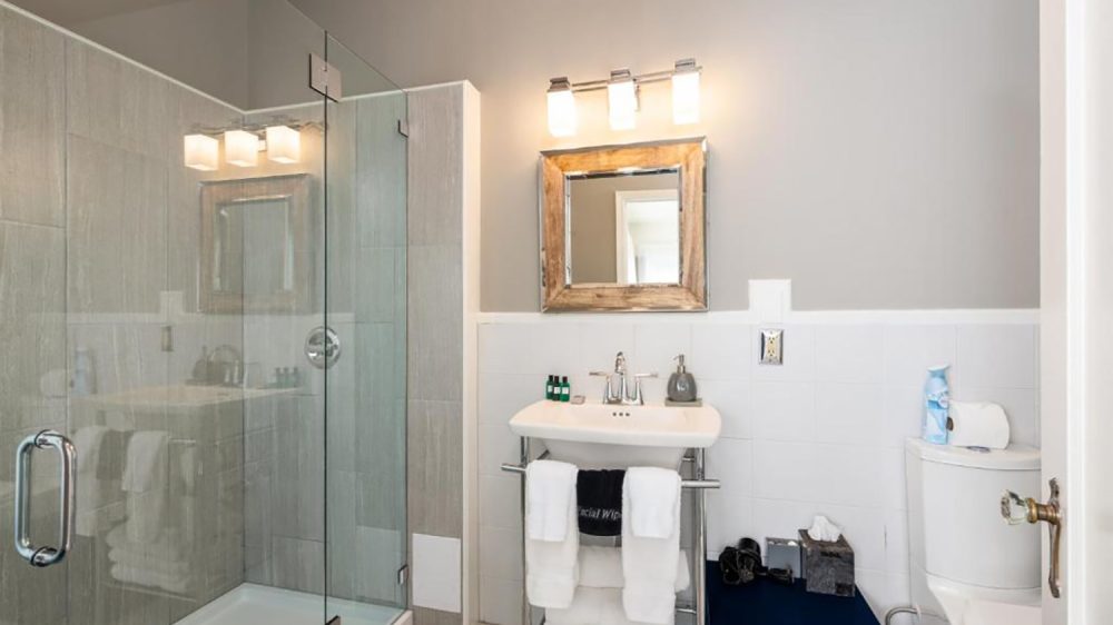 Modern bathroom with a glass-enclosed shower featuring gray tiles, a white pedestal sink with chrome fixtures and towels hanging on a rack beneath it, a wooden-framed mirror above the sink, and a white toilet with a blue shelf beside it holding tissue and air freshener. The walls are painted light gray with white tile halfway up.