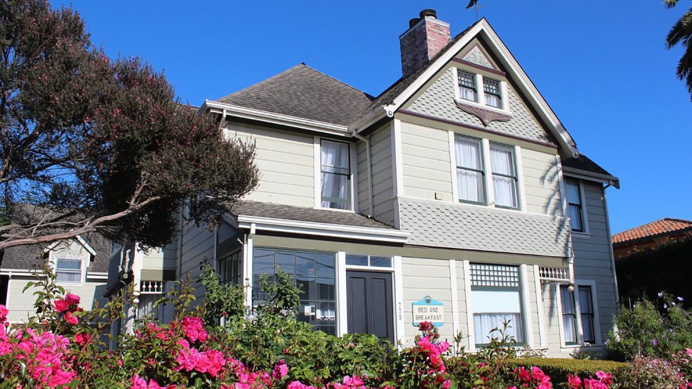 Two-story bed and breakfast house with light gray siding, a dark gray shingled roof, and a brick chimney, surrounded by vibrant pink flowers and greenery under a clear blue sky.