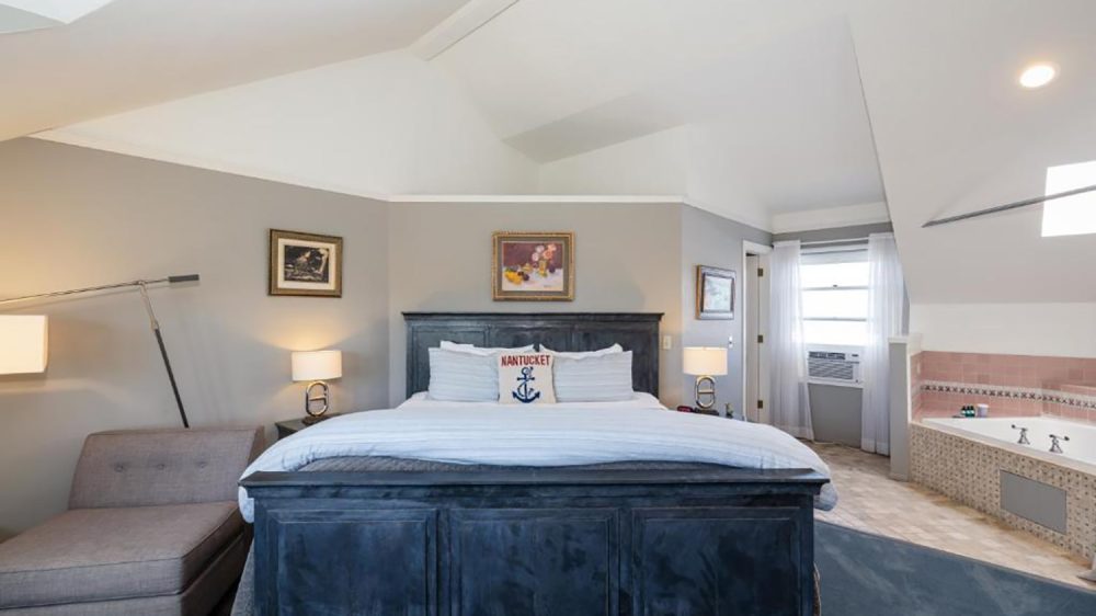 Bedroom with a large dark wooden bed featuring white and striped bedding and a decorative pillow with "Nantucket" and an anchor design. The room has gray walls, two bedside tables with lamps, a gray chaise lounge on the left, and framed artwork above the bed. To the right, there is a window with sheer white curtains and an air conditioning unit, and a tiled bathtub with pink and beige tiles. The ceiling is vaulted and painted white.