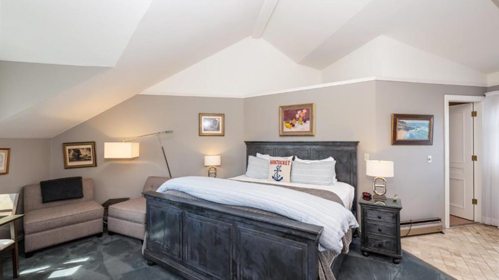 Bedroom with a dark wooden bed frame and white bedding, featuring a decorative pillow with "Nantucket" and an anchor design. The room has gray walls and a sloped ceiling. There are two beige upholstered chairs with a black throw blanket on one, a floor lamp with a rectangular shade, and two bedside tables with lamps. Several framed paintings hang on the walls. The floor is carpeted in a dark gray color, and there is a doorway leading to another room with tiled flooring.