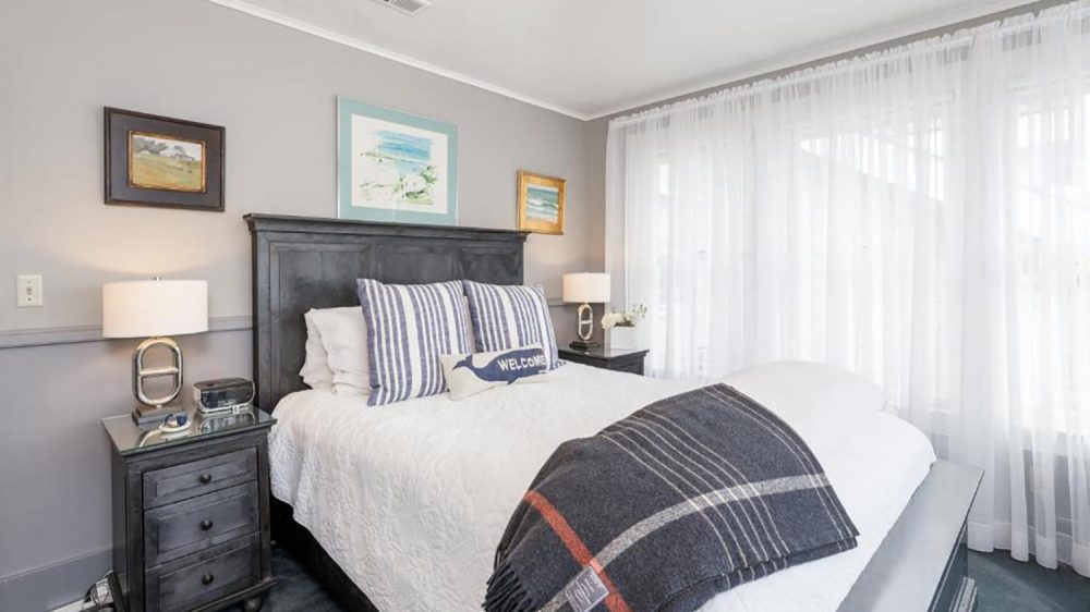 Bed with a dark wooden headboard and matching nightstands on either side, each with a lamp. The bed is made with white bedding, two striped pillows, and a small rectangular pillow with a whale and the word "WELCOME." A dark plaid throw blanket is draped across the foot of the bed. Light gray walls with three framed paintings hang above the headboard. Sheer white curtains cover the window on the right side.