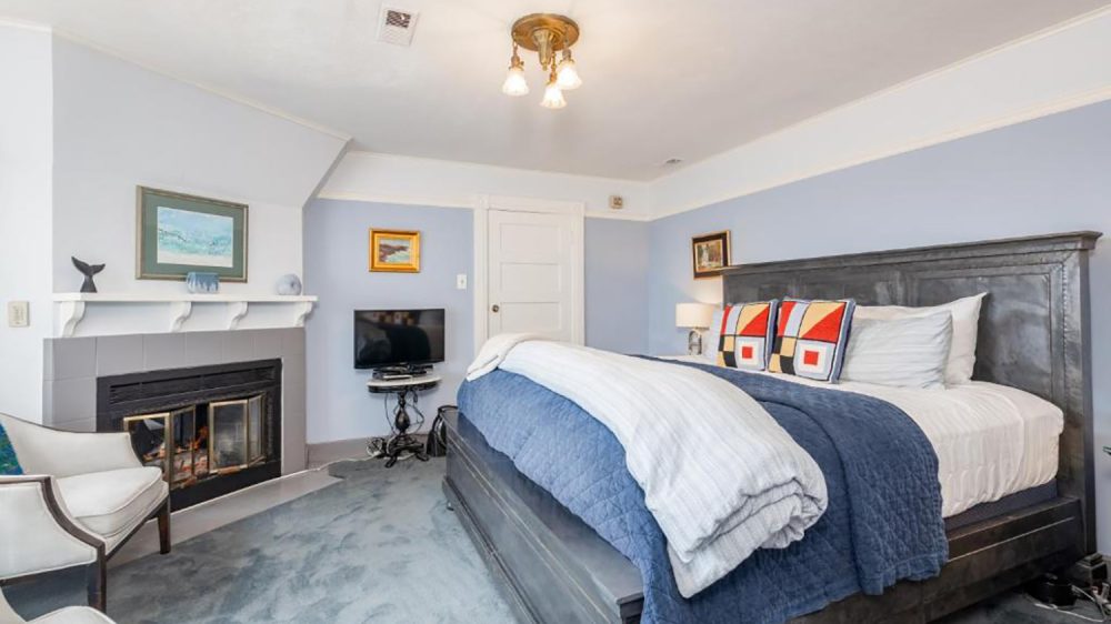 Bedroom with a large dark wood bed featuring a blue quilt and white bedding, accented by three colorful geometric pillows. The room has light blue walls, a gray carpet, and a white ceiling with a brass light fixture. There is a fireplace with a white mantel decorated with small sculptures and a framed picture above it. A small TV sits on a black ornate stand near a white door. Two white armchairs are positioned near the fireplace.
