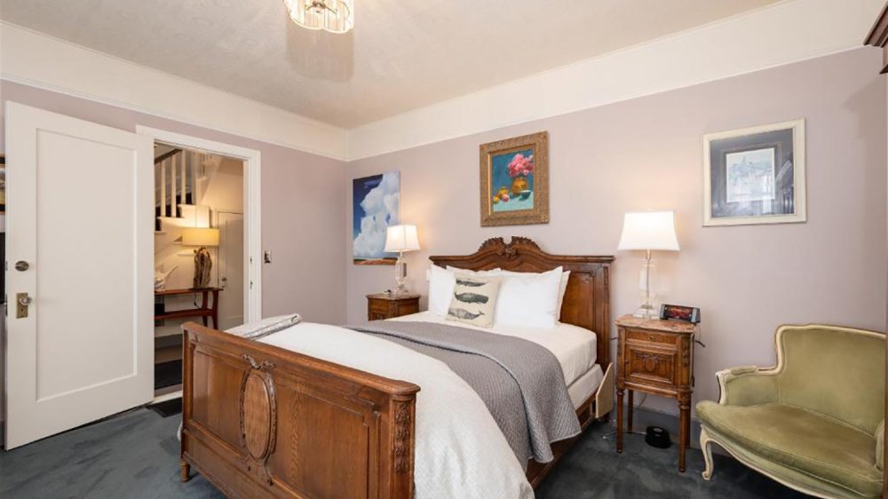 Bedroom with a wooden bed frame featuring intricate carvings, white bedding, and a gray throw blanket. Two wooden nightstands with lamps flank the bed. The walls are painted light lavender with three framed artworks hanging above the bed and on the side wall. A green upholstered loveseat is positioned to the right of the bed. The room has a blue carpet and a white ceiling with a decorative light fixture. An open door reveals a hallway with a staircase and a table lamp.