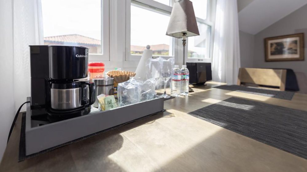 A wooden table with a black coffee maker, coffee supplies including filters and sugar packets, two water bottles, two wine glasses, a paper towel roll, a lamp with a beige shade, and a black speaker. The table is near large windows with white curtains, and there are black placemats on the table. A framed picture is visible on the wall in the background.