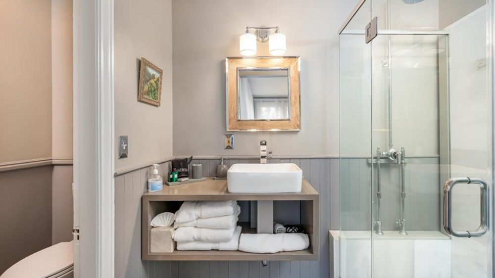 Modern bathroom with a floating vanity featuring a rectangular white vessel sink, a large square mirror with a wooden frame, and a double light fixture above. The vanity shelf holds neatly folded white towels and a tissue box. To the right, there is a glass-enclosed shower with a chrome rain showerhead and fixtures. The walls are painted in neutral tones with wainscoting, and a small framed painting hangs on the left wall near the toilet.