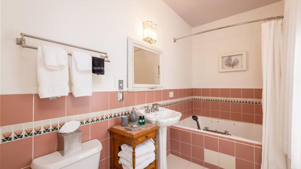 Bathroom with pink and white tiled walls, featuring a white pedestal sink, a bathtub with a handheld showerhead, a white toilet, and a wooden cabinet holding folded white towels. A white-framed mirror and a wall light are above the sink, with white towels hanging on a metal towel rack. A white shower curtain is partially drawn around the bathtub.