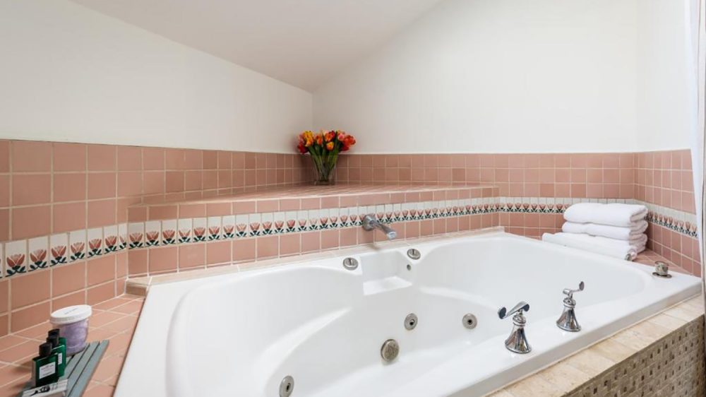 White corner jacuzzi bathtub with silver faucets, surrounded by pink tiles featuring a floral border. A vase of orange and red flowers is placed on the tiled ledge behind the tub, with neatly folded white towels stacked on the right side. Small bottles and a container are arranged on the left side ledge. The walls above the tiles are plain white.