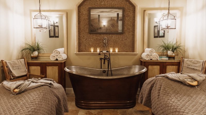 A cozy spa room featuring a dark bronze freestanding bathtub centered between two wooden cabinets with glass tops. Each cabinet holds a stack of rolled white towels and a potted green plant. Above each cabinet is a large rectangular mirror, and hanging pendant lights with multiple bulbs illuminate the space. Two massage tables with quilted gray covers and robes are positioned on either side of the tub. The wall behind the tub has a mosaic tile inset with a seahorse decoration, five lit candles, and a framed beach pathway photograph. The overall color scheme is warm and neutral.