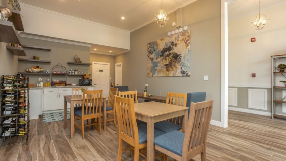 Dining area with wooden tables and chairs featuring blue cushions, light wood flooring, and neutral-colored walls. A large abstract painting with tree motifs hangs on one wall. The room includes a small kitchenette with white cabinets, open shelves holding various items, a microwave, and a wine rack. Two spherical pendant lights hang from the ceiling, and an adjacent room is visible through a large doorway.