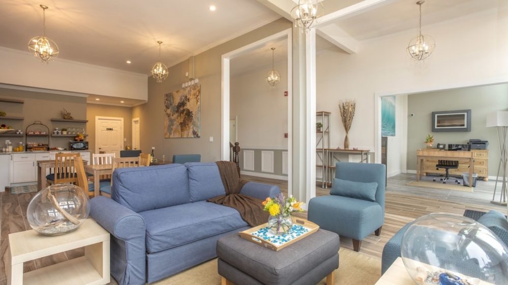 A modern living room with a blue sofa, matching blue armchairs, and a dark gray ottoman with a wooden tray holding a vase of flowers. Light wood side tables with decorative glass bowls flank the seating. The room features beige walls, wood flooring, and three spherical pendant lights. In the background, there is a dining area with a wooden table and chairs, a kitchen counter with shelves holding various items, and an adjacent office space with a wooden desk and chair.