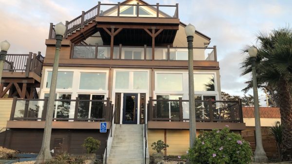 A multi-story building with large glass windows and brown wooden railings on the balconies. There is a central staircase leading up to a dark-colored door. Four tall street lamps are positioned in front of the building, with plants and shrubs around the base. The sky is partly cloudy with soft lighting.