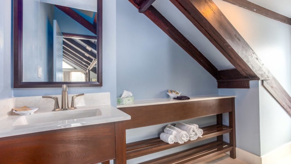 Bathroom vanity area with a white countertop and a silver faucet, a large dark-framed mirror above the sink, and a long wooden shelf underneath a slanted ceiling with exposed dark wooden beams. Rolled white towels are neatly placed on the lower shelf. The walls are painted light blue.