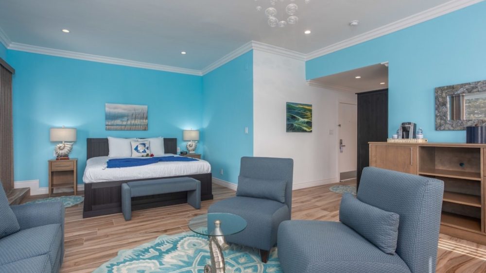 Bedroom with bright blue walls featuring a dark wood bed with white bedding and blue accents, two matching nightstands with shell-themed lamps, and a blue patterned rug. The seating area includes two blue upholstered chairs and a glass-top side table with a seahorse base. A wooden cabinet with a coffee maker and water bottles is against one wall, and there are two framed artworks and a mirror on the walls. The floor is light wood, and the ceiling has recessed lighting and a bubble-style chandelier.