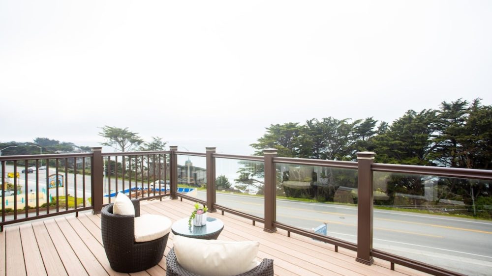 Balcony with light wood flooring and dark brown wicker chairs with white cushions, accompanied by a small round glass-top table with a small plant. The balcony has a railing with glass panels and overlooks a road, trees, and a distant view of the ocean.