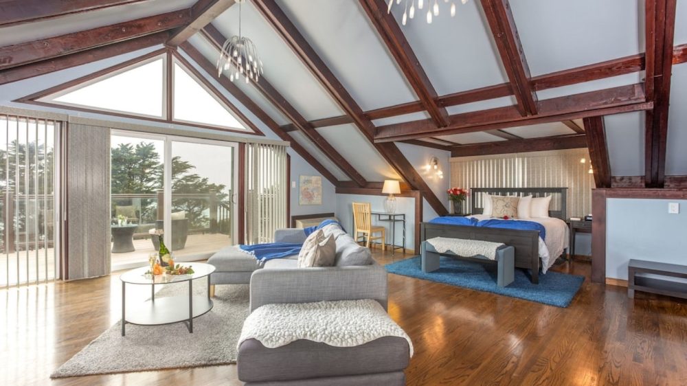 Spacious attic bedroom with exposed dark wooden beams and light blue walls. The room features a large bed with white and blue bedding on a blue rug, a gray sectional sofa with a white throw, and a round glass coffee table on a beige rug. A wooden chair and small desk with a lamp are positioned near the bed. Large sliding glass doors with vertical blinds open to a balcony with outdoor seating, allowing natural light to fill the room. Modern pendant lights hang from the ceiling. The floor is polished hardwood.