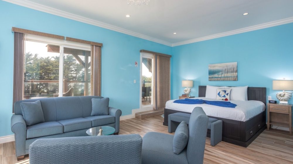 Bedroom with light blue walls and wood flooring featuring a dark wood king-size bed with white bedding and a blue throw. Two matching nightstands with lamps flank the bed. A blue sofa and two blue armchairs surround a small glass coffee table. Large sliding glass doors with vertical blinds lead to an outdoor area with trees visible outside. A coastal-themed painting hangs above the bed.