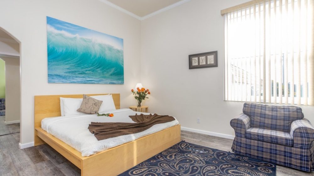 Bedroom with a light wooden bed frame and white bedding, featuring a brown throw and a decorative pillow. A small wooden nightstand with a lamp and a vase of orange roses is beside the bed. A large wall art of a blue ocean wave hangs above the bed. The floor is gray wood, partially covered by a blue patterned rug. A blue plaid armchair sits near a large window with vertical blinds. The walls are painted white.