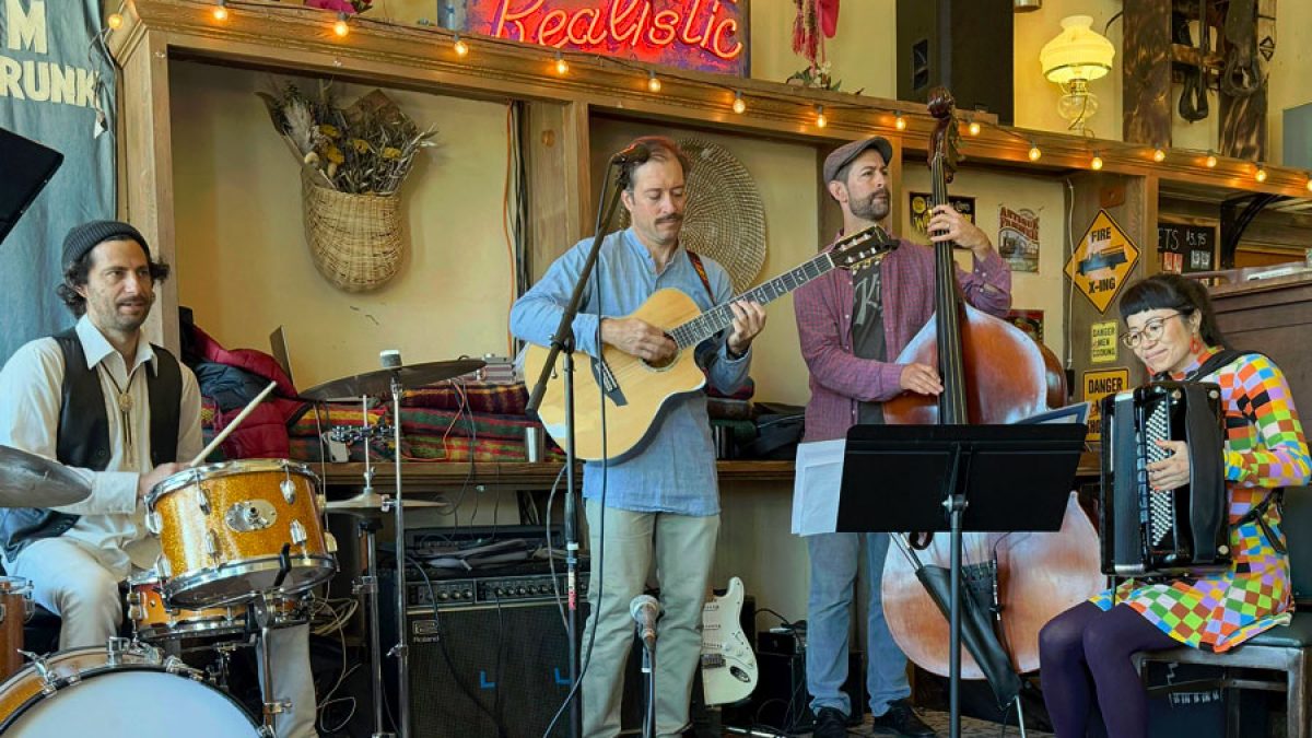 A four-member band performing indoors with a drummer playing a gold drum set, a guitarist in a blue shirt, a double bass player wearing a cap and plaid jacket, and a woman in a colorful checkered dress playing an accordion. The background features a shelf with decorative items, a neon sign that reads "Realistic," and various wall signs.