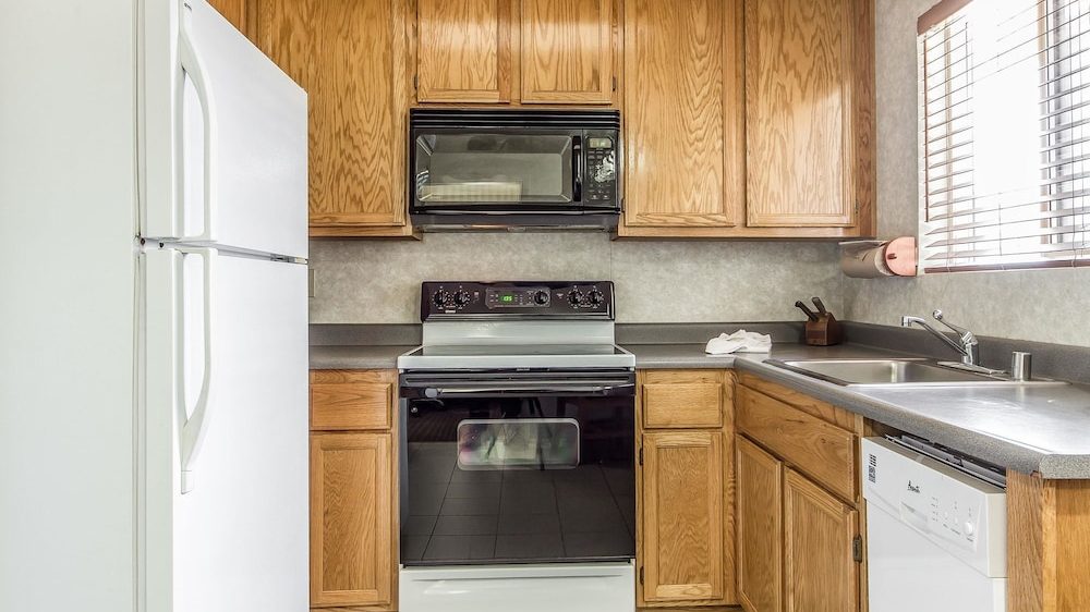Kitchen with wooden cabinets, a white refrigerator on the left, a black and white electric stove with an oven in the center, a black microwave above the stove, and a double stainless steel sink with a faucet on the right. There is a dishwasher below the counter next to the sink and a window with blinds above the sink. The countertops are gray.