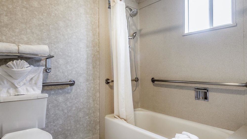 Bathroom with a white bathtub and shower combination, featuring a white shower curtain. There are stainless steel grab bars along the walls near the bathtub. A small window above the bathtub allows natural light in. To the left, a toilet with a metal rack above it holds neatly folded white towels, including one folded in a decorative fan shape. The walls have a textured, light gray finish, and the floor is tiled in white.