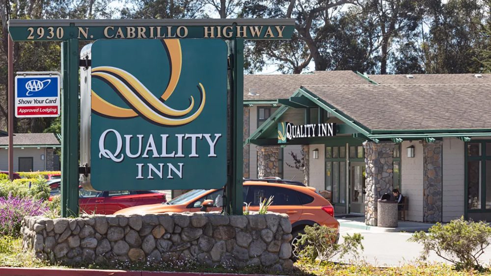 Sign for Quality Inn at 2930 N. Cabrillo Highway, featuring a green background with yellow and white swoosh design, mounted on a stone base. In the background, a building with stone pillars and green trim is visible, along with parked cars and trees.