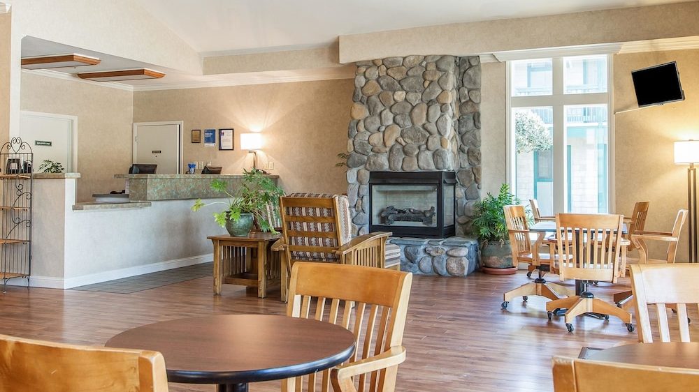 Lobby area with wooden tables and chairs, a stone fireplace, potted plants, a reception desk, and a wall-mounted TV near large windows.