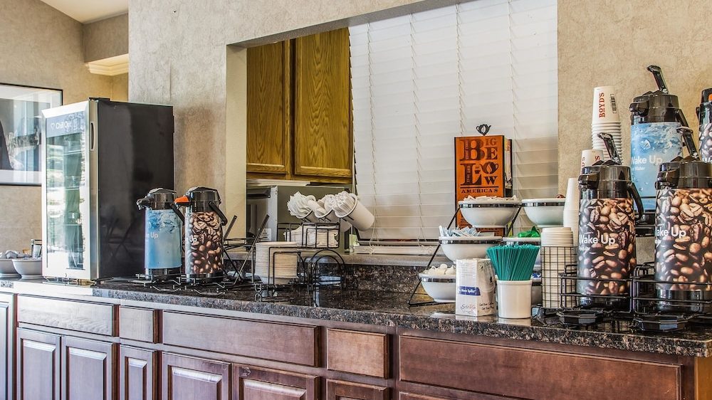 A coffee station setup on a dark granite countertop with wooden cabinets below. The station includes multiple airpots with coffee bean designs labeled "Wake Up," a small refrigerator, stacks of disposable cups, green stirrers in a white container, sugar packets, and bowls with creamers or sweeteners. There is a window with closed white blinds and a framed picture on the wall to the left.