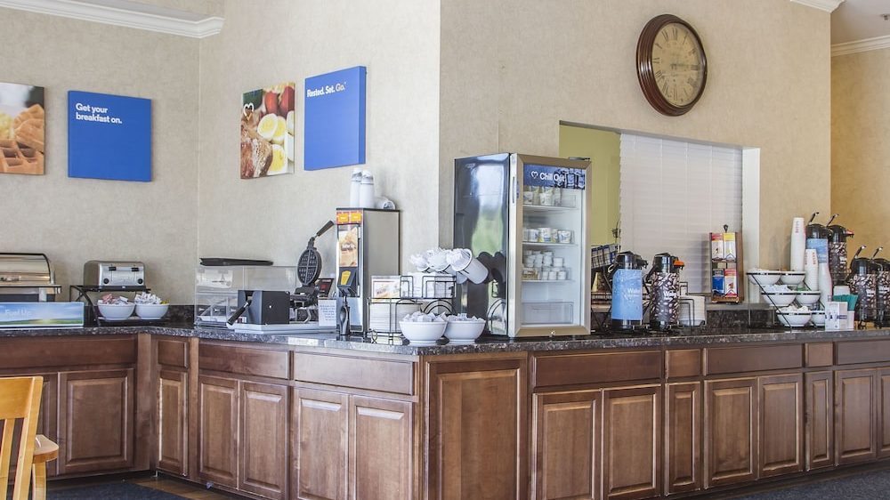Breakfast buffet area with wooden cabinets, a waffle maker, bowls of cereal, a small refrigerator stocked with yogurt, coffee dispensers, and a wall clock above a pass-through window. Blue signs on the wall read "Get your breakfast on." and "Rested. Set. Go.