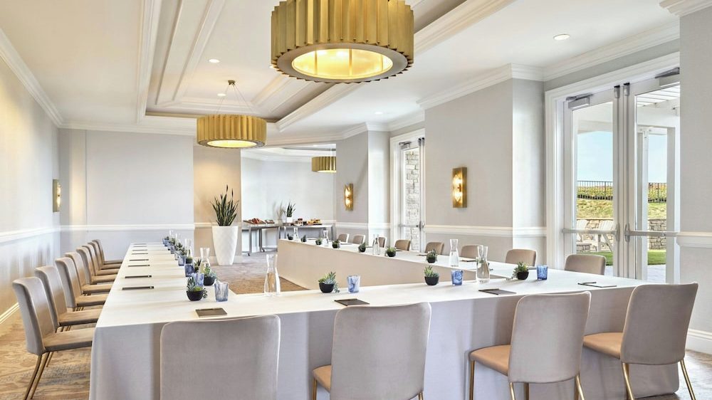 A bright conference room with a U-shaped table covered in white tablecloths, surrounded by beige upholstered chairs with gold legs. The table is set with small potted plants, blue glasses, water carafes, and notepads. The room features large windows and glass doors letting in natural light, light gray walls with white trim, and three large, gold, drum-shaped pendant lights hanging from the ceiling. A buffet table with refreshments is visible in the background.