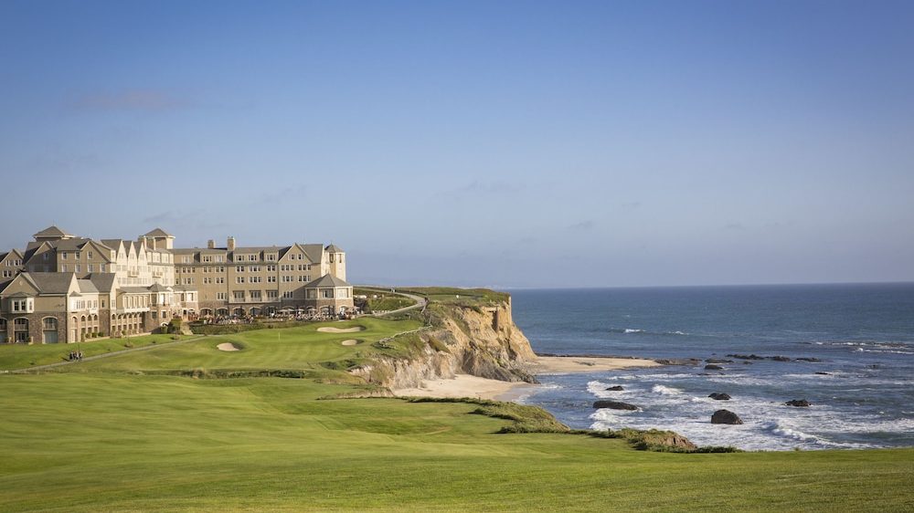 A large beige stone building, likely a hotel or resort, sits on a grassy cliff overlooking a rocky shoreline and the ocean under a clear blue sky. The foreground features a well-maintained green lawn, possibly part of a golf course.