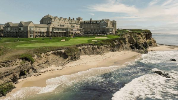 A large, multi-story stone building with a complex roofline sits atop a grassy cliff overlooking a sandy beach and ocean waves. The building appears to be a hotel or resort, with green lawns and golf course features in front. The sky is partly cloudy with soft light.