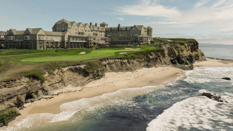 A large, multi-story stone building with a complex roofline sits atop a grassy cliff overlooking a sandy beach and ocean waves. The building appears to be a hotel or resort, with green lawns and golf course features in front. The sky is partly cloudy with soft light.