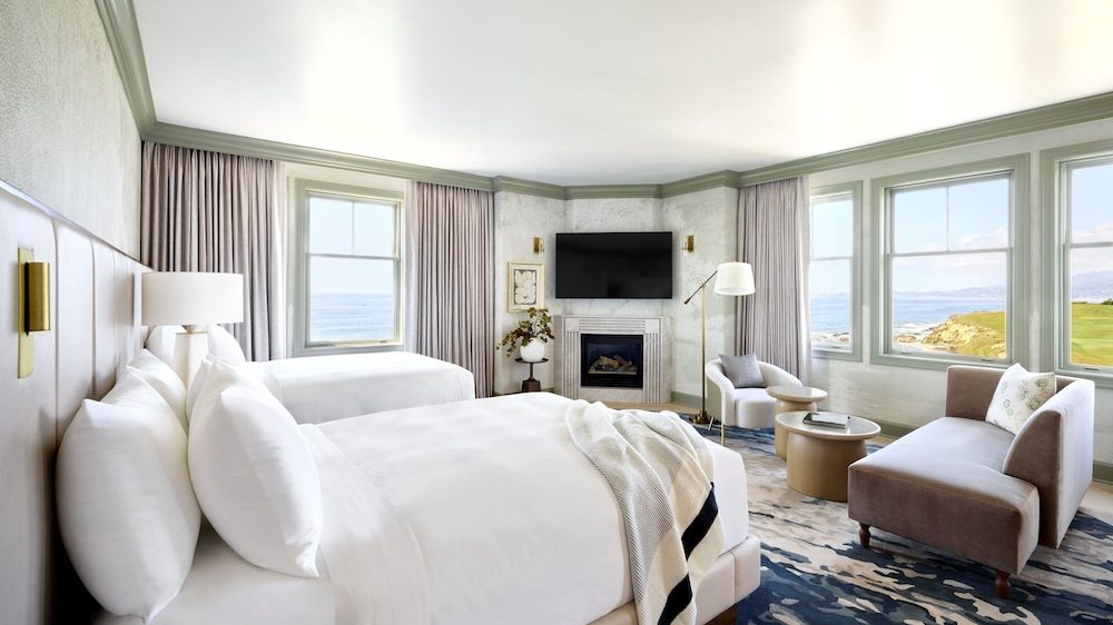A bright, spacious hotel room featuring two large beds with white linens and multiple pillows. The room includes a cozy sitting area with a beige loveseat, a white armchair, and two round coffee tables. Large windows offer a scenic view of the ocean and coastline. A flat-screen TV is mounted above a modern fireplace, and the room is decorated with neutral tones, soft curtains, and a patterned blue and beige rug.